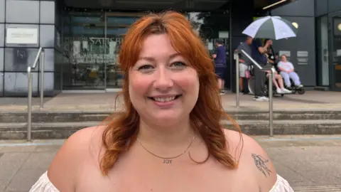A young woman with ginger hair is smiling. She wears a white top and a necklace which says "JOY".
