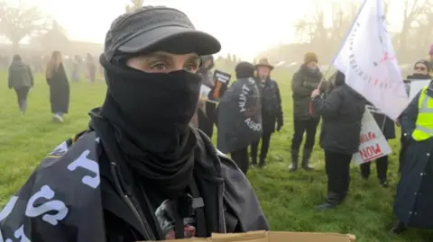 In the foreground is a person wearing a black cap, and a black face covering and only their eyes are visible. They are draped in a flag and wearing black clothing. They are looking just off camera. Behind them are a few peope gathered waving flags and holding placquards. They are all in a field which a layer of light fog.