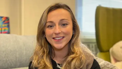 Charlie is sitting on a light grey sofa in a bright room with large windows. She is wearing a black top and a sleeveless brown cardigan. Behind her, a colourful piece of artwork rests on the windowsill, and soft furnishings are visible on the right.