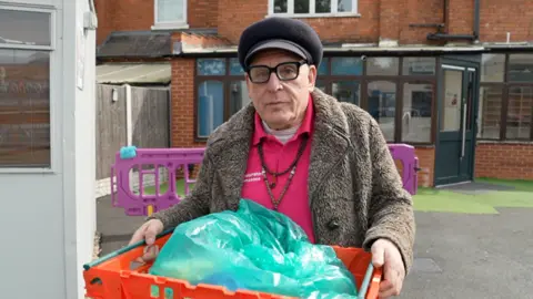 Volunteer driver Pete Hammond carries an orange box containing a green bag of donated food. He is wearing thick, black rimmed glasses and a black Rasta-style hat and the pink embroidered polo shirt bearing the food bank's logo with a fake fur coat on top. He has necklaces showing over his pink polo shirt.