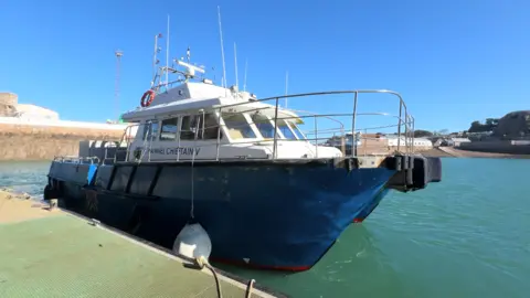 A blue and white vessel named Channel Chieftain V moored at Victoria Harbour. The boat has a sturdy metal rail around the deck, various antennas and equipment on the roof, and a white fender hanging from the side. It sits in calm turquoise water, with a harbour wall and nearby buildings visible in the background under clear blue sky.