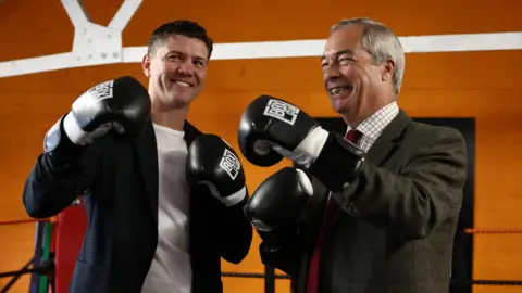 EPA Luke Campbell and Nigel Farage stand in a boxing ring wearing black boxing gloves. Campbell, on the left of the image, is smiling as he wears a dark jacket and white t-shirt. Farage, on the right, is wearing a green tweed jacket, shirt and maroon tie.