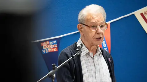 PA Media A man in a grey shirt and blue cardigan, he has white hair and glasses and is giving a speech in front of a microphone