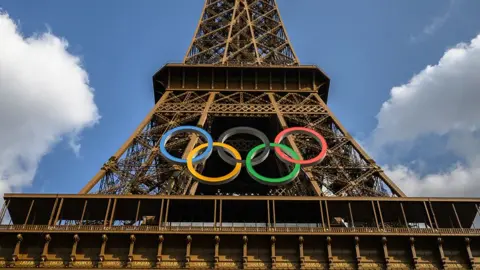 The olympic rings logo are seen attached to the front of the Eiffel tower on a sunny day with some clouds in the background, in Paris on 24 July