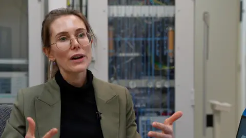 A woman in a khaki jacket, black polo neck jumper and round glasses talks to a camera off centre, gesticulating with her hands. She is sitting in a lab with various bits of equipment out of vision behind her. she has dark hair tied back.