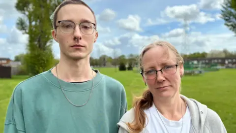 On the left is a young man with cropped dark hair, glasses and a nose ring, wearing a green sweatshirt. On the right is a woman with long light brown hair tied in a pony tail and glasses. She's wearing a white t shirt and grey jacket. They are standing in a park and the grass, blue and sky and clouds in the background are blurred.