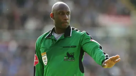 Uriah Rennie, the first black referee in the Premier League, is pictured holding his hand out while wearing a green shirt refereeing a game.