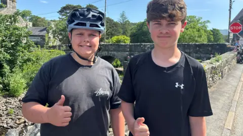Two teenage boys looking at the camera. One is wearing a bike helmet and a grey top. He is smiling at the camera. The other is wearing a black top with an under armour logo. He is brown hair.