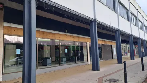 A vacant retail unit on a parade of shops. Structural pillars hold the upper floor of the parade aloft.