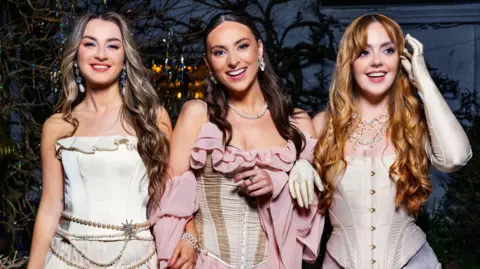 Three women wearing dresses and jewellery. They are standing with their arms linked and smiling.