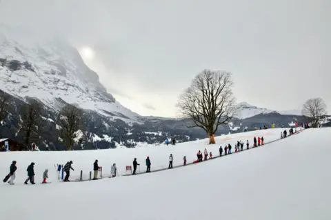 Pemain ski Thierry Meyrat di sabuk konveyor di Grindelwald, Swiss. Para pemain ski berada di garis menuju lereng. Ada gunung yang tertutup salju di belakang mereka.