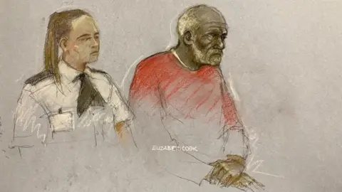 Elizabeth Cook/PA Media Ryland Headley wearing a red jumper in a court sketch. He is sitting in the dock next to a female security officer. He has his palms crossed on his lap in front of him.