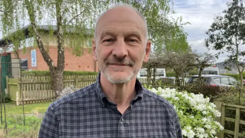 A man is smiling at the camera. He has grey hair and a grey beard. He is wearing a grey checked shirt and standing in a garden. 