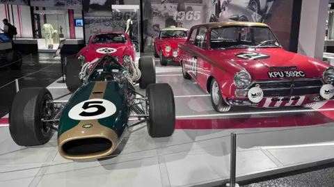 Kate Bradbrook/BBC A dark green and gold racing car with a number 5 on the front. There is a red car behind it and two further red cars to the right. One has the number 27 in white writing on the bonnet. They are parked on a grey stage at an exhibition.