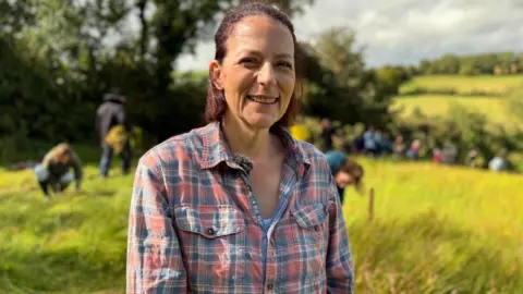 Helen has short hair. She's wearing a checked top. She's standing in a field of flax. People are working in the field behind her.