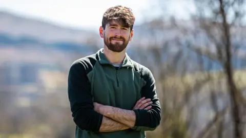 Rhys Evans, director of the Nature Friendly Farming Network in Wales, standing in a rural area, with his arms folded and smiling at the camera