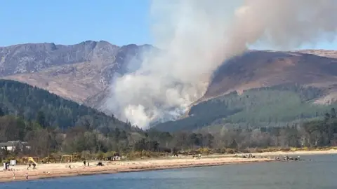 A wide shot of a large fire in the hills above a beach in Arran. Smoke can be seen rising from the grassy slope.