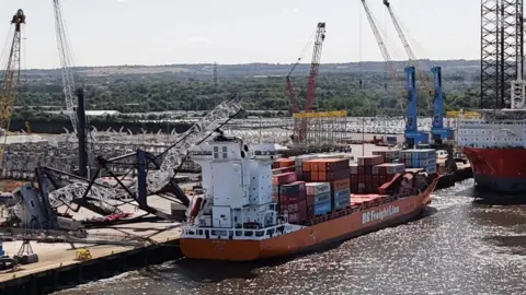 Aerial view of a container ship at a dock. It is a low-lying, orange coloured vessel, its deck stacked with containers. A crane lies crumpled next to it with the top part resting on the vessel's control tower.