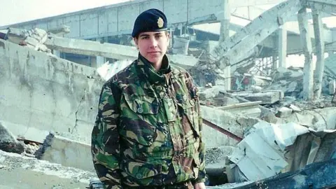 Jemma Morgan Jemma Morgan wore camouflage uniforms and a black military cap. She stands between debris on active duty and a destroyed building. She is looking at the camera with an empty expression