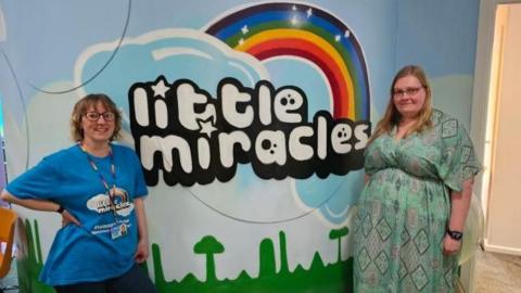 Two women stand in front of a Little Miracles poster 