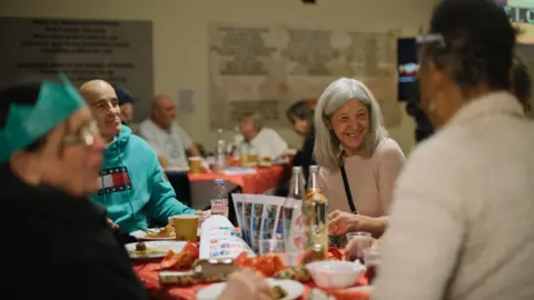 The Salvation Army People sat around a table which has Christmas crackers on and decorations. One woman with grey hair and wearing light pink top is smiling at another woman who has her back to the camera. 