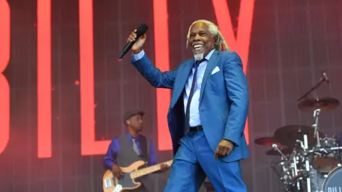 Jim Dyson/Getty Images A singer wearing a blue suit smiling while standing onstage holding aloft a microphone.