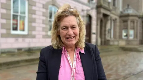 BBC Andy Howell pictured in the Royal Square. The states assembly building is visible behind her. She is wearing a pink shirt and black jacket. She has blonde shoulder length wavy hair and a pair of reading glasses on her head. She is wearing a Government of Jersey lanyard. 