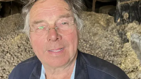 John Devine/BBC Brian Croft is standing in front of a barn which is full of potatoes. Brian has short grey hair and is he is wearing a black jacket and is wearing clear framed glasses.