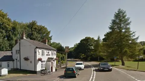 Google Deanshanger village, with a traditional white pub on the left and cars parked alongside. Another car is heading towards the camera.  A village green is visible to the right.