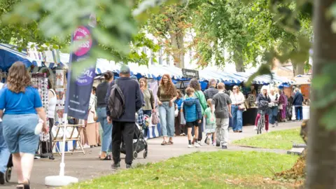 A busy outdoor market with the leaves of trees hanging over the top of the photo. People are busy shopping and walking through 