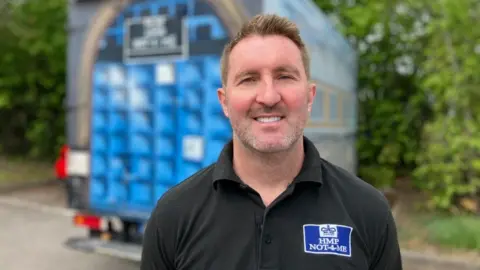 A middle-age white man with brown hair wearing a black polo neck T-shirt with a mock prison logo on the chest. He is standing in front of a bright-blue van with prison artwork painted on the sides. He looks relaxed and is smiling directly at the camera.