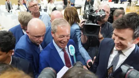 PA Media A tightly-packed crowd of men, some holding cameras. The none at the centre has grey hair. He is wearing a blue suit and shirt with a red tie and blue and white rosette and is smiliing.