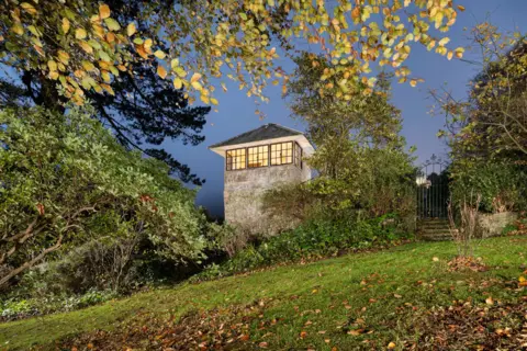 Historic England Archive The summerhouse at Upper Cobb House is a tall rectangular granite tower which has windows wrapped around its top storey under a steep slate roof. The windows are lit. There is a wrought iron gate to the right and grass, shrubs and beech trees in the foreground.