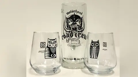 Eddisons Three ale glasses on a shelf. One has a cat image, another an owl and a tall one in the centre has the words Motorhead and road crew.