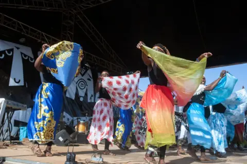 Amos Gumulira / AFP A group of female refugees perform on stage, holding up material that matches their skirts as part of a  traditional cultural dance at the Dzaleka Refugee Camp in Dowa, Malawi - Saturday 2 November 2024