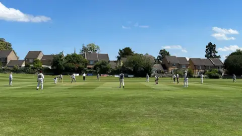 Tom Williams/BBC A wide view of people playing cricket on a field on a bright and sunny day. There is a row of houses at the back of the field. 