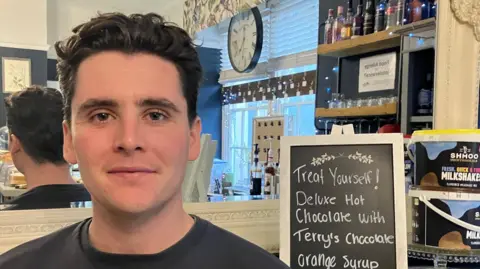  Gareth, a young man with short dark hair sitting in a cafe. Beside him is a chalk board advertising deluxe hot cholocate.