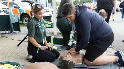 A Gorman Photography A man wearing a hooded jumper performs compressions on a medical dummy whilst being watched by a female paramedic at the Bath and West Show