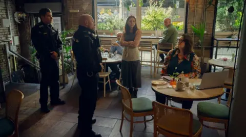 Run Away Private investigator Elena Ravenscroft, played by Ruth Jones, is sitting on a table in a restaurant and talking to two police officers. A number of customers who are sitting at different tables are staring in their direction. 