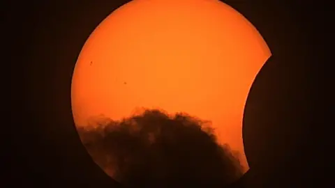 BBC Weather Watchers/The Happy Wanderer The partial eclipse captured in Blyth. The sun appears like a large orange circle, with a small chunk missing from the right hand side. There is a cloud passing at the bottom of the sun