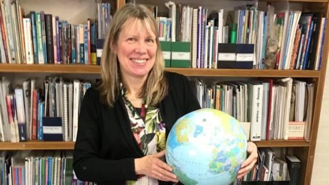 Wendy Shimmin, wearing a black jacket, holds a globe in front a bookcase