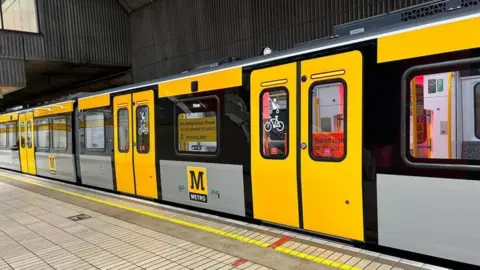 A Metro train pulling into Gateshead Interchange. The train is yellow and silver and displays the Metro M logo. The station floor is made of beige tiles.