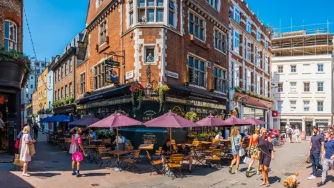 Busy London street with people dining outside Shakespeare’s Head pub under umbrellas, surrounded by historic buildings and shops on a sunny day.