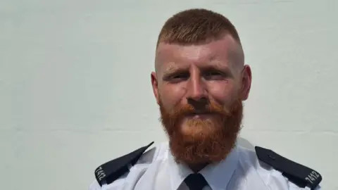 A man in a white and black police uniform standing in front of a blank, white wall.