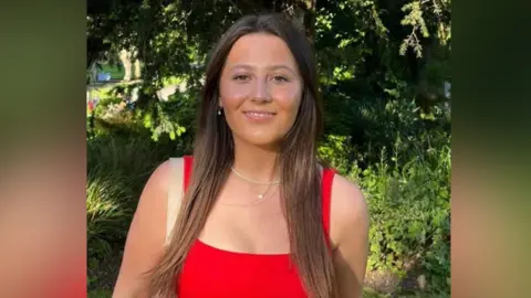 Supplied A young woman with a bright red top on. Behind her are leafy trees and shrubs. 