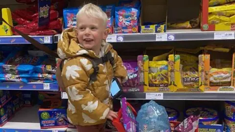 Family Handout Oscar wearing a mustard coat pushing a child's shopping trolley with sweets in the basket in Asda.