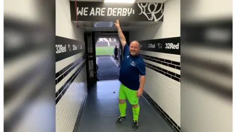 SUPPLIED Bald man with navy football shirt and neon green football shorts and socks pointing to a Derby County Football Club sign saying 'we are Derby' in a corridor leading to a football pitch in the background