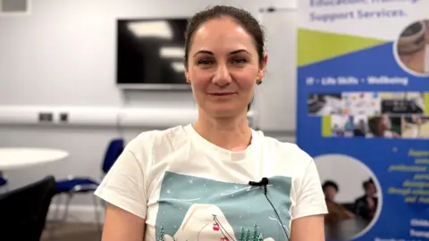 Alesea has brown hair pulled back, she is earing a white T-shirt and on it is an artwork of a ski slope with red cable cars making their way upwards.