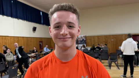 Jeff is wearing an orange top and has light brown lifted-up hair. He is standing in a school assembly hall.