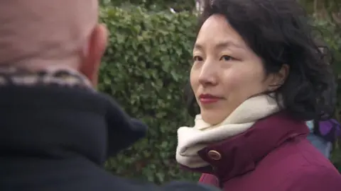 MP Yuan Yang standing outside the school wearing a maroon coat and a white scarf. She is looking at a parent who is talking at the protest. 